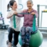A care provider helps an older woman exercise with a resistance band and an exercise ball, representing how exercise can help with senior fall prevention.